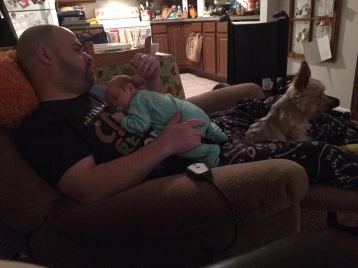 Photo of dad holding baby and small dog, watching television. Baby is sleeping.